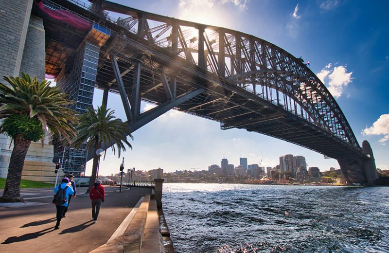 People walking along the Sydney Harbour waterfront under the Sydney Harbour Bridge on a sunny day