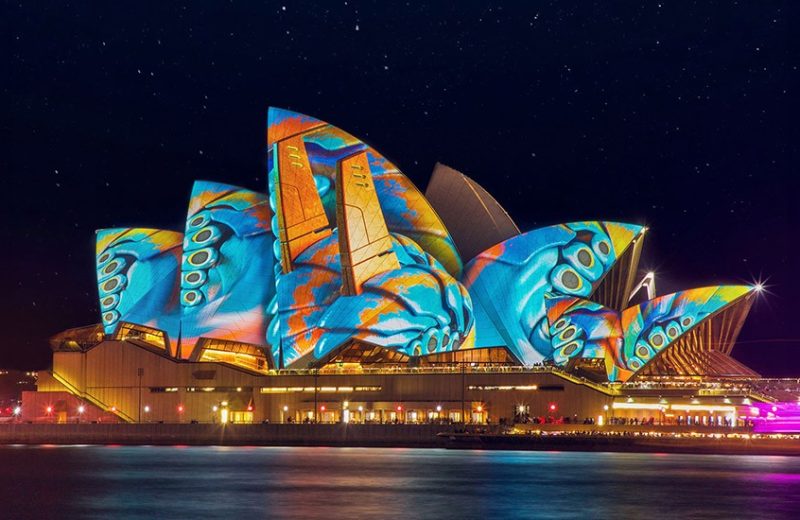 A vibrant nighttime photo of the Sydney Opera House sails lit up with colourful projections, reflecting on the harbour water during Vivid Sydney