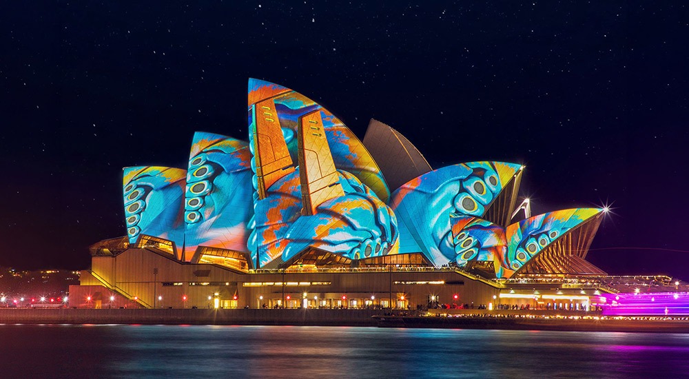 A vibrant nighttime photo of the Sydney Opera House sails lit up with colourful projections, reflecting on the harbour water during Vivid Sydney